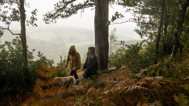 Two people walking a dog in the rain. They are walking along a wooded path with a misty view behind them